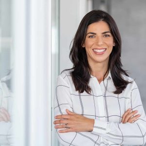 Portrait of middle aged businesswoman in modern office looking at camera. Confident business woman with arms crossed standing while leaning against glass wall. Proud brunette woman smiling in formalwear with copy space.